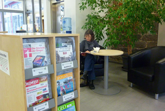 Eine Frau sitzt lesend in einer Sitzecke mit Sesseln und Tisch. Im Vordergrund: Regale mit Zeitschriften zum Ausleihen.
