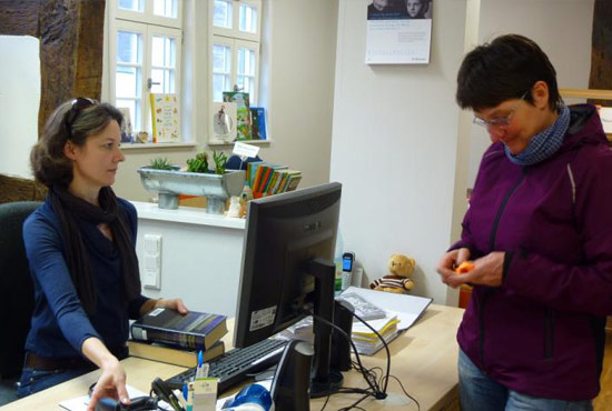 Ausleihvorgang. Bibliotheksmitarbeiterin und Bibliotheksnutzerin, dazwischen ein Tisch mit Rechner. Die Nutzerin leiht mehrere Bücher. Die Bibliotheksmitarbeiterin erfasst die Bücher im System.