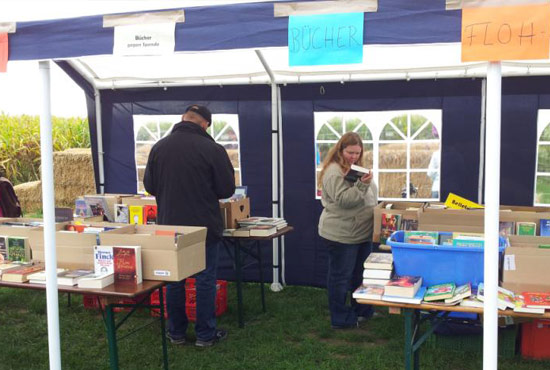 Bücherflohmarkt: Vor einem Maisfeld auf einer Wiese unter freiem Himmel aufgebautes Zelt mit Tischen, auf denen Bücher zum Verkauf angeboten werden. Ein Herr und eine Dame stöbern in den Büchern.