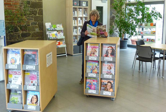 Rollbare halbhohe offene Schränke mit Zeitschriften in weitläufigem Raum. Eine Frau steht an einen der Rollschränke angelehnt und schmökert in einer Zeitschrift.