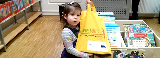 Lesestart: dreijähriges Mädchen mit Lesestart-Tasche vor Bilderbüchern